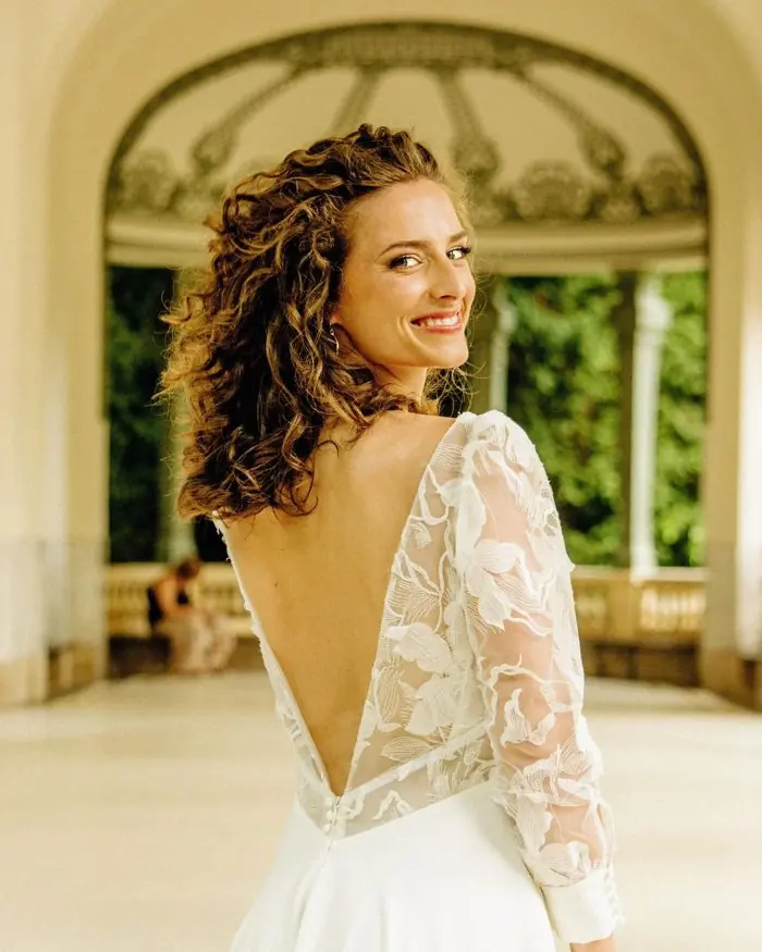 Mariée en robe à dos dentelle, cheveux bouclés, souriante par-dessus l'épaule